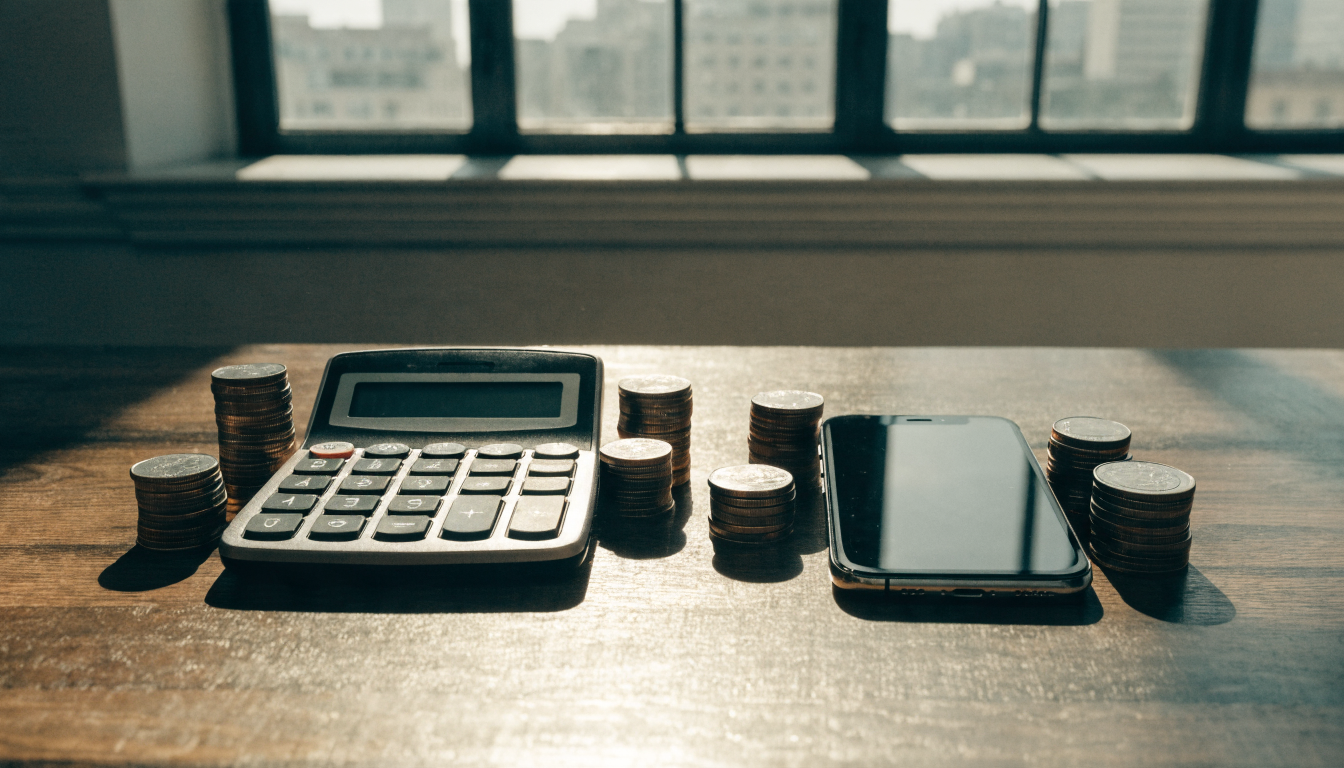A calculator and a smartphone side by side on a desk, with small stacks of coins representing subscription costs